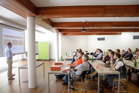 Mann hält vor einem Smartboard einen Vortrag vor einer Gruppe von Menschen